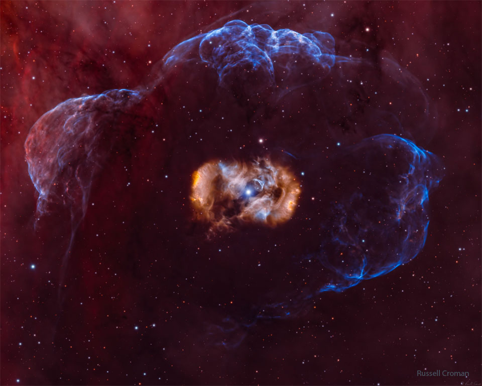 这张特色图片显示一颗恒星位于一个对称但复杂的多色星云内，星云周围环绕着一个淡蓝色星云。有关更多详细信息，请参阅说明。