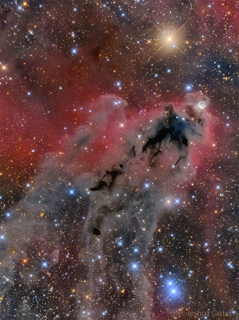 一张不祥的黑暗星云的图像，背景是红色发光的气体和许多明亮多彩的恒星。有关更多详细信息，请参阅说明。