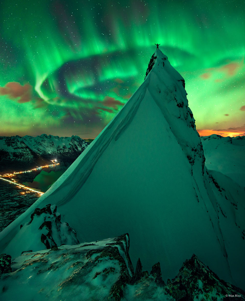 一个人站在陡峭的白雪覆盖的小山上，双臂高举。远处可见绿色的极光。透过极光可见群星闪烁。有关更多详细信息，请参阅说明。