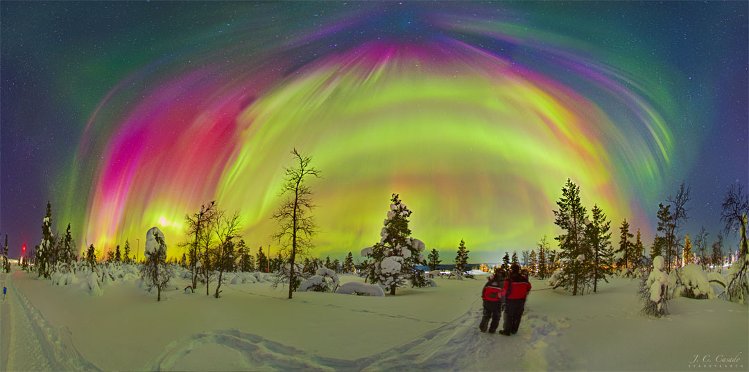 两个穿着红色外套的人站在一片白雪皑皑、树木裸露的土地上。上图中，出现了许多不同颜色的极光，背景中可以看到一些恒星。有关更多详细信息，请参阅解释。