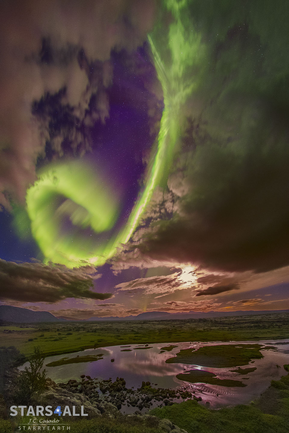 在紫色背景前的云层间隙中可以看到亮绿色的螺旋极光。前景是绿色的草原和一个圆形的湖泊。有关更多详细信息，请参阅说明。