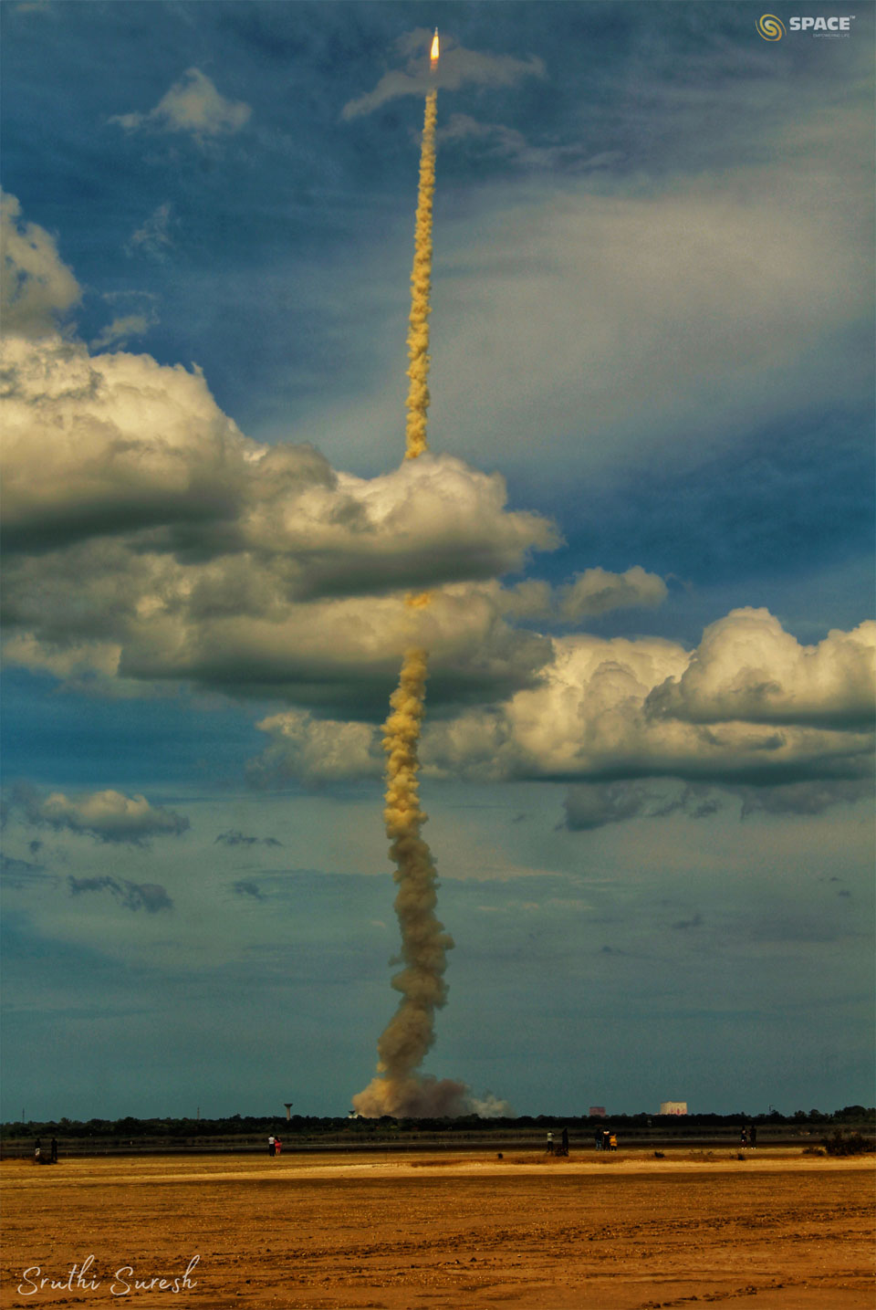 火箭升空后冒出长长的烟羽。火箭在蓝天的映衬下穿过云层。前景是一片空旷的棕褐色田野。有关更多详细信息，请参阅说明。
