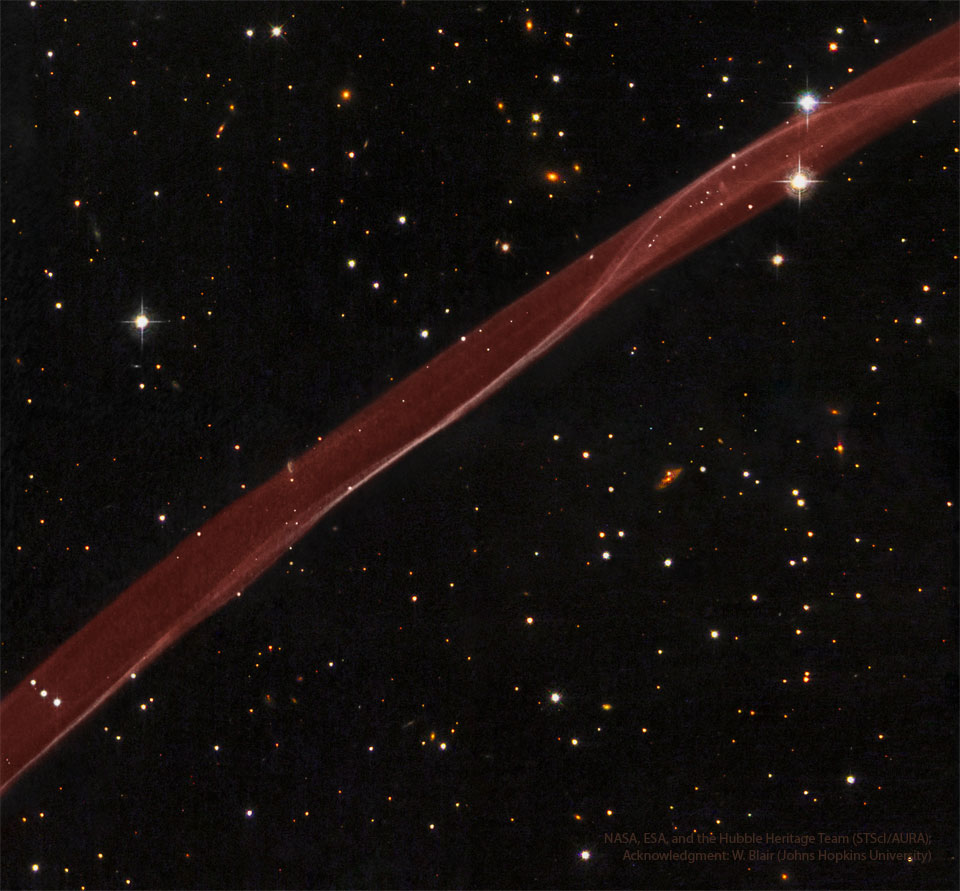 一条厚厚的透明红色气体带从左下角流向右上角。明亮的红色气体带周围是一片布满星星和星系的黑暗星域。有关更多详细信息，请参阅说明。
