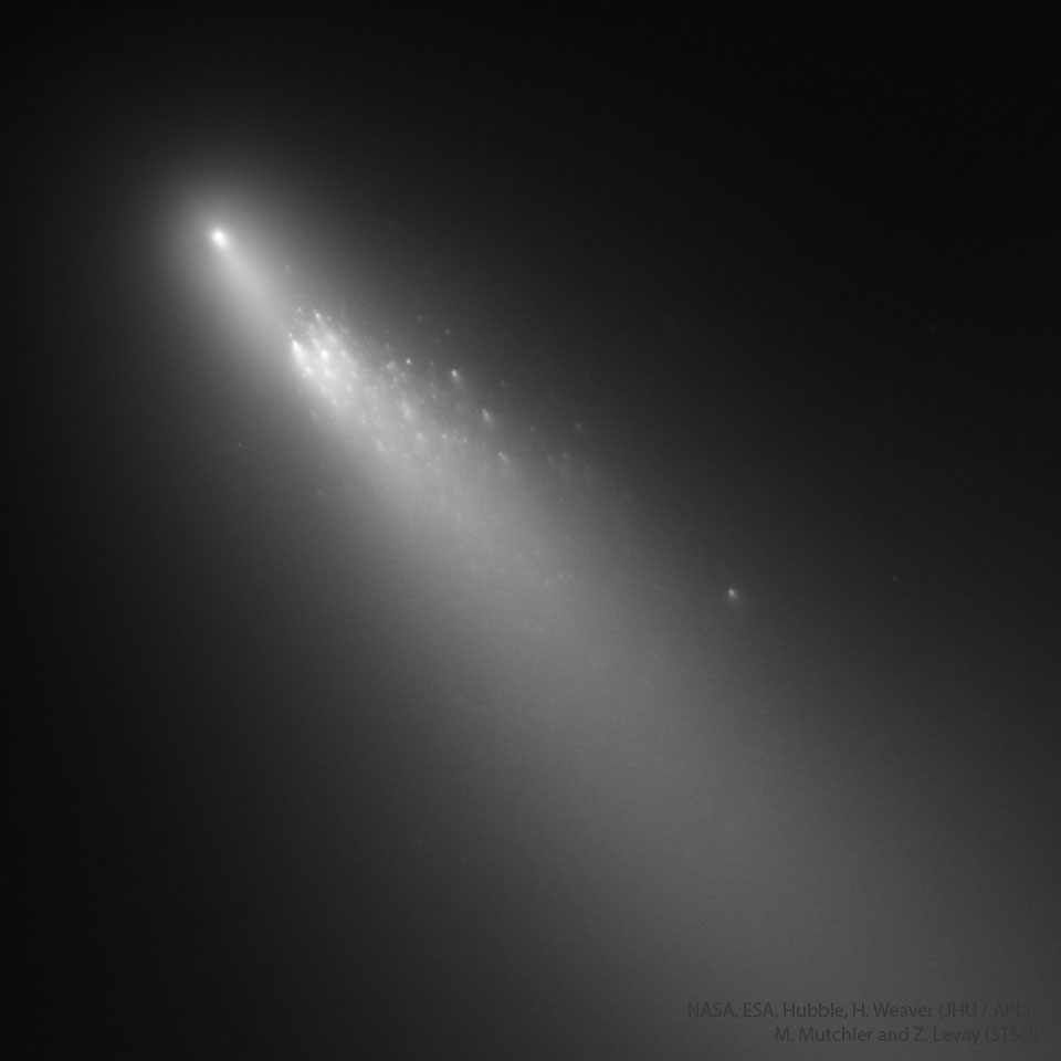 在黑暗的太空背景下，一颗模糊的彗星以灰色显示在左上角。彗星的尾巴呈对角线延伸到右下角。彗星的主要部分被分解成许多碎片。有关更多详细信息，请参阅说明。