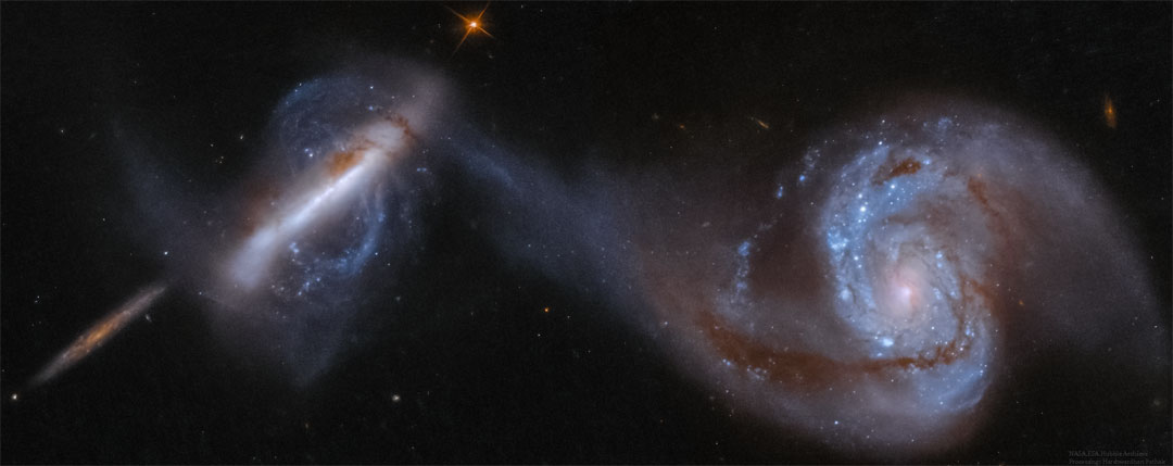 图中显示了三个大星系，最右边的两个正在碰撞。最右边的星系是一个巨大的螺旋星系，它的一只臂与左边一个不寻常的极地星系相连。最左边较小的星系被认为远在背景中。有关更多详细信息，请参阅说明。