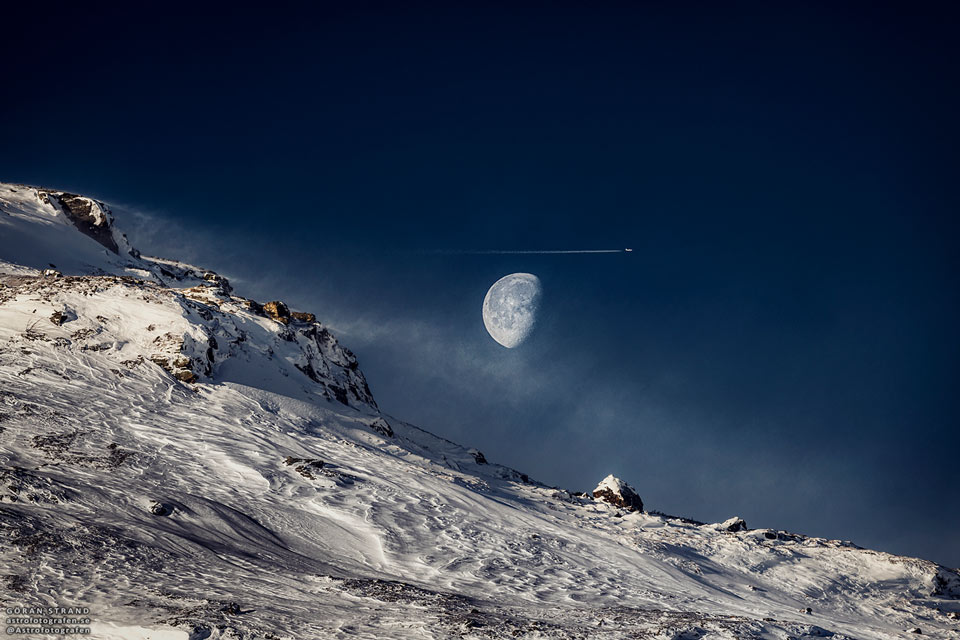 在白雪皑皑的斜坡上可以看到一轮满月。在月亮附近可以看到一架飞机和尾迹。有关更多详细信息，请参阅说明。