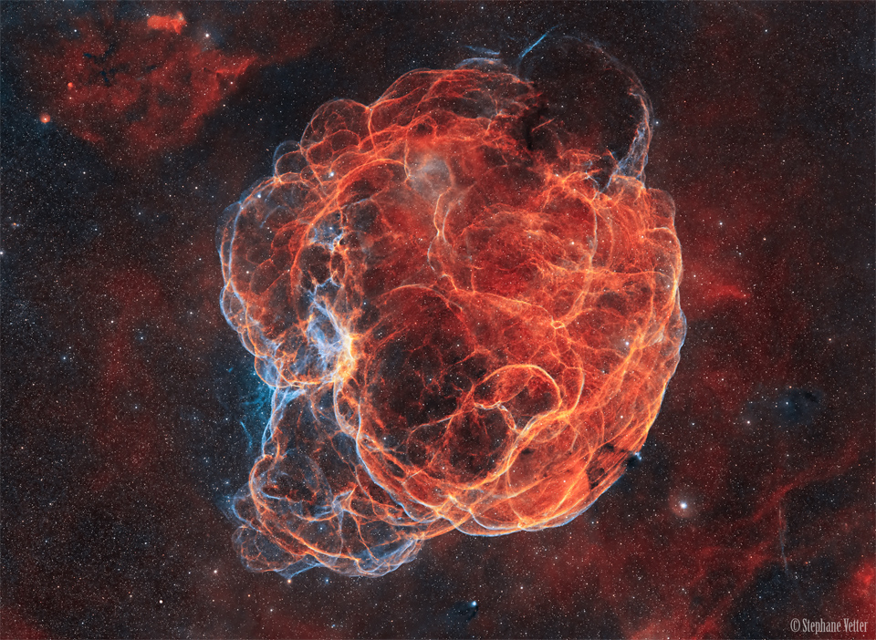 一个巨大的丝状星云被红色的光所占据，但在左下方有一些蓝色的光。该星云位于密集的星场中，周围环绕着其他微弱的红色发光星云。有关更多详细信息，请参阅说明。
