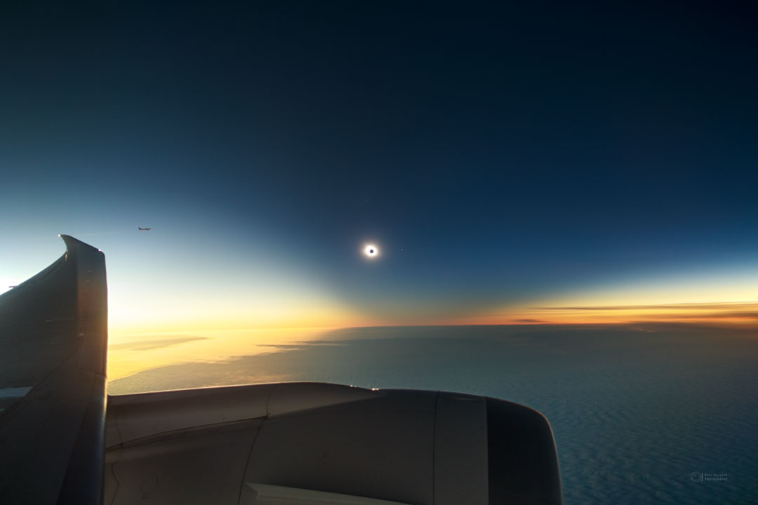 远处可以看到一个日全食的太阳。日食周围是一个从上方向下倾斜的黑暗区域。下面是云层，再下面是飞机的机翼和发动机。有关更多详细信息，请参阅说明。
