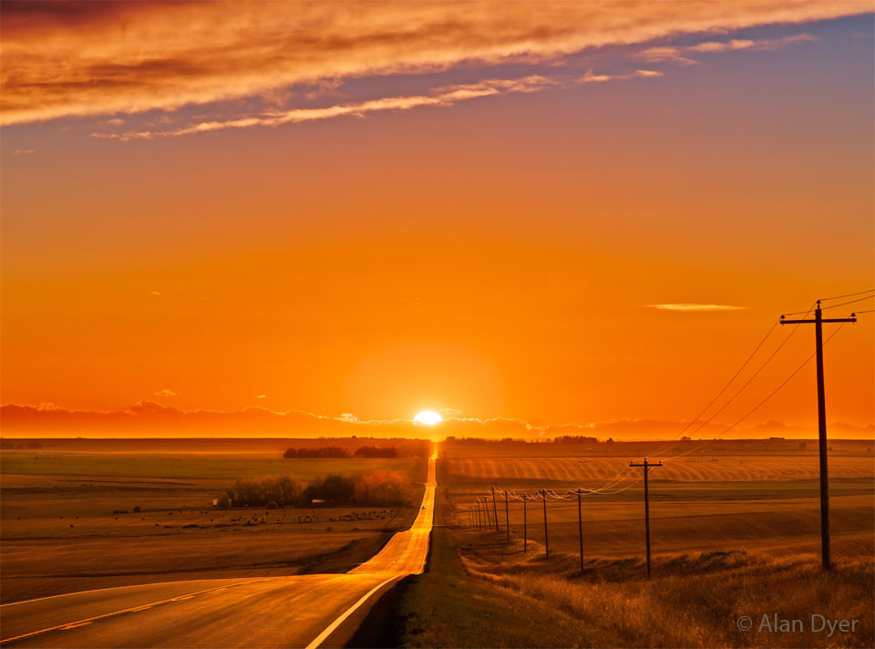 太阳在遥远的地平线上落下，在开阔的田野上一条长长的道路的尽头。日落是橙色的，就像周围的天空一样。电线杆排列在路的右侧。有关更多详细信息，请参阅说明。